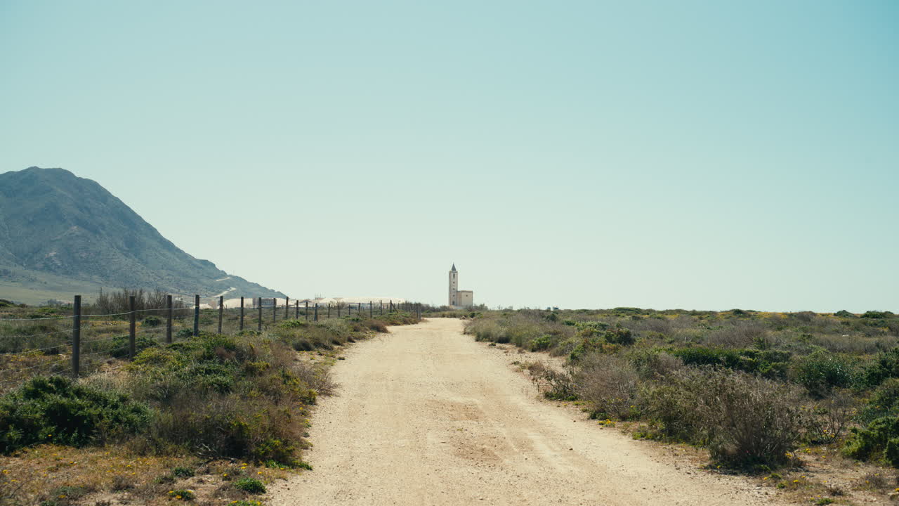 Pintoresco camino de tierra que se extiende a través de un paisaje rural seco hacia una torre de iglesia distante, con montañas y cielo azul claro. Fuertes líneas de fuga y espacio abierto crean una sensación de viaje y aventura.