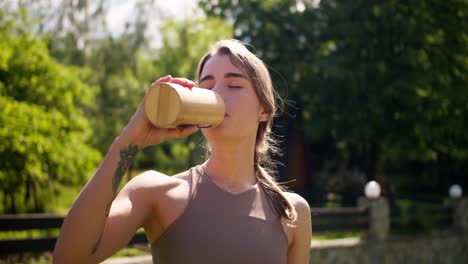 a young blonde girl in a special summer sports uniform drinks water from a yellow bottle against the backdrop of a green forest