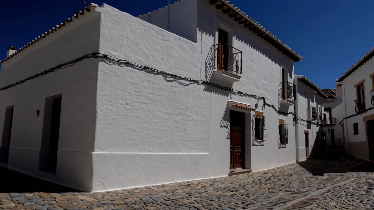 Traditional whitewashed houses with balconies and barred windows line a narrow cobblestone street, creating a picturesque scene in an Andalusian village under the vibrant blue sky
