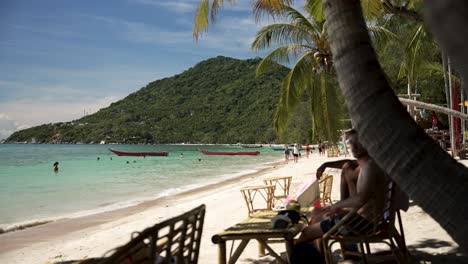 A scenic view of Sairee Beach, Koh Tao, Thailand, featuring tourists relaxing on the sandy shore under palm trees and enjoying the turquoise waters.
