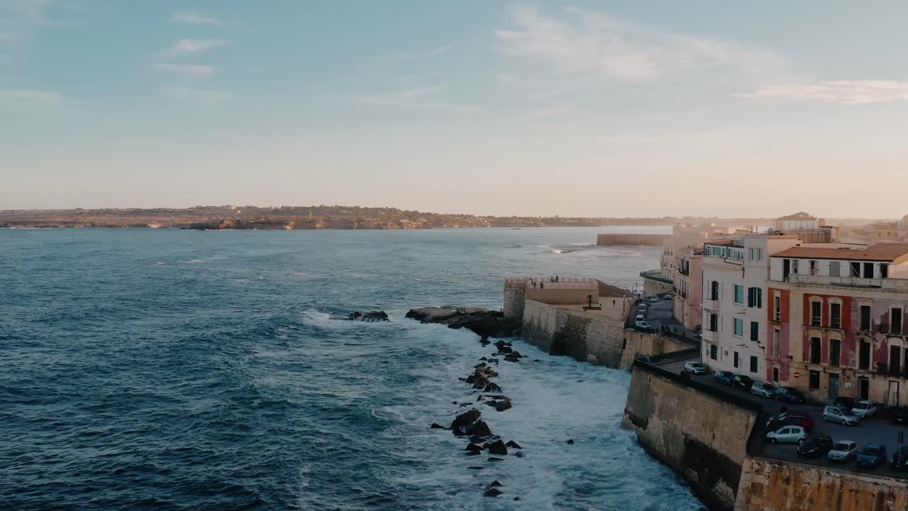 Aerial drone shot of Ortigia island along the Mediterranean sea cost with large waves crashing on the fortress. Historic charming medieval town in Sicily at sunset.