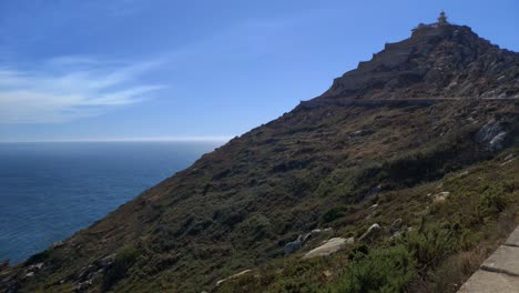 Path-along-the-cliff-of-the-mountain-to-the-summit-with-viewpoint-and-lighthouse-in-the-Atlantic-ocean,-sunny-day,-panoramic-shot-turning-right,-Cíes-Islands,-Pontevedra,-Galicia,-Spain