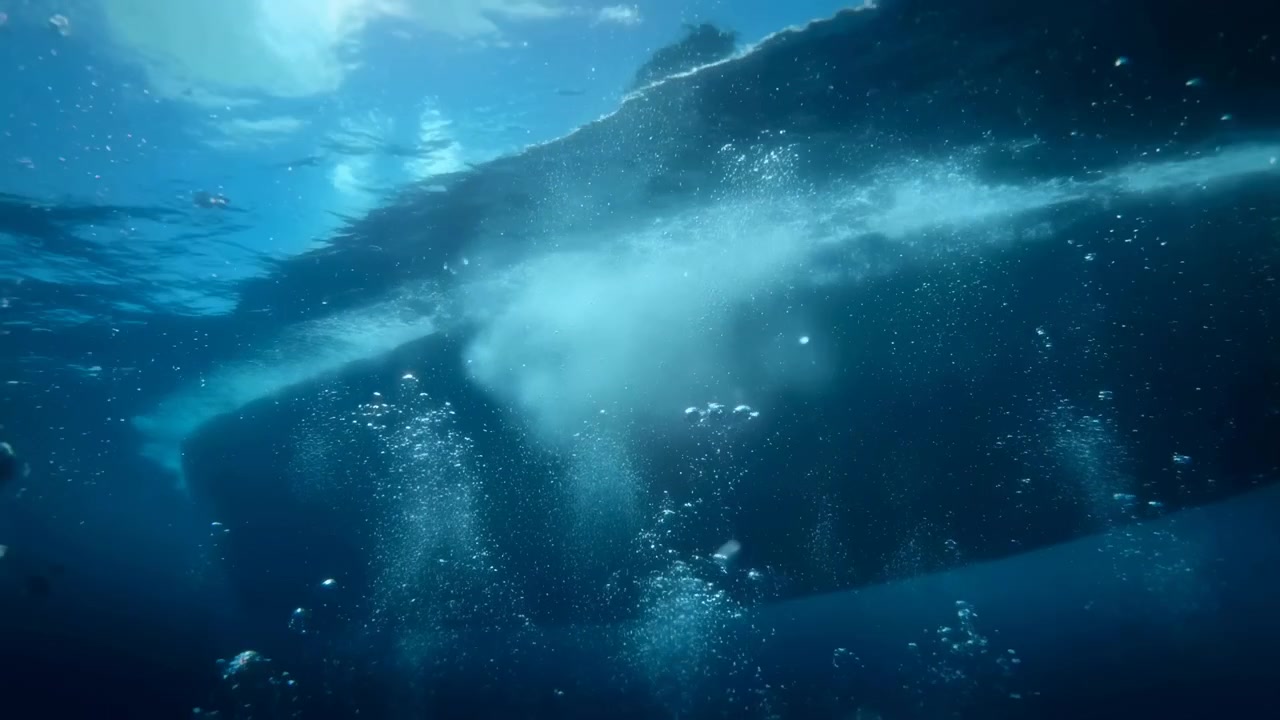Seen from below the surface, a powerful wave passes across the frame while bright foam disperses and clusters of bubbles rise through dark blue water under shifting natural light.