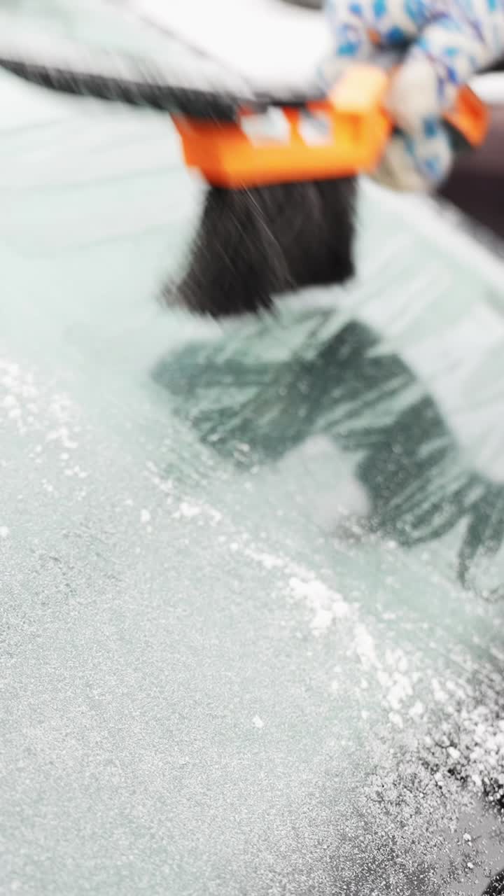 Gloved hand sweeps a snow brush across an icy windshield, removing frost and loose snow to restore visibility before driving. Vertical close-up winter b-roll