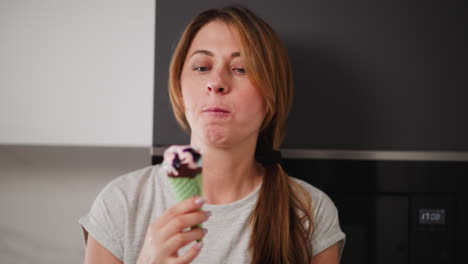 portrait view of lady with long hair wearing casual grey shirt enjoying colorful ice cream cone with green wafer in bright modern kitchen, smiling subtly while savoring sweet treat