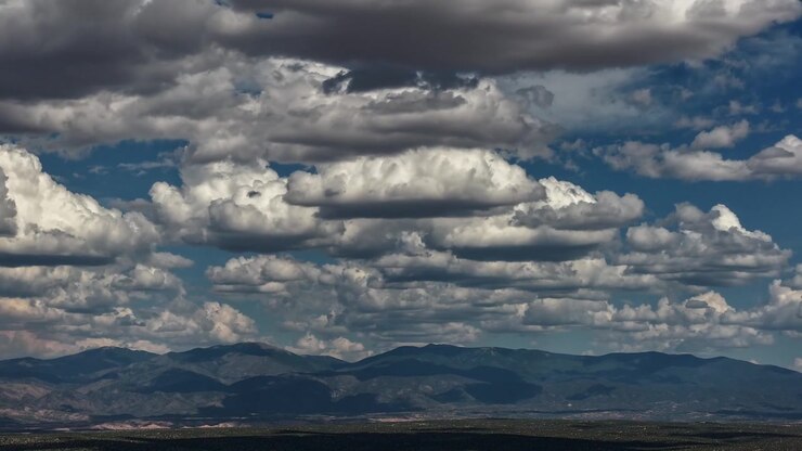 4K drone time lapse capturing dramatic, multi-tiered layers of white and grey clouds shifting across a blue sky. Deep shadows crawl across Mt Baldy and the vast Santa Fe County desert below.