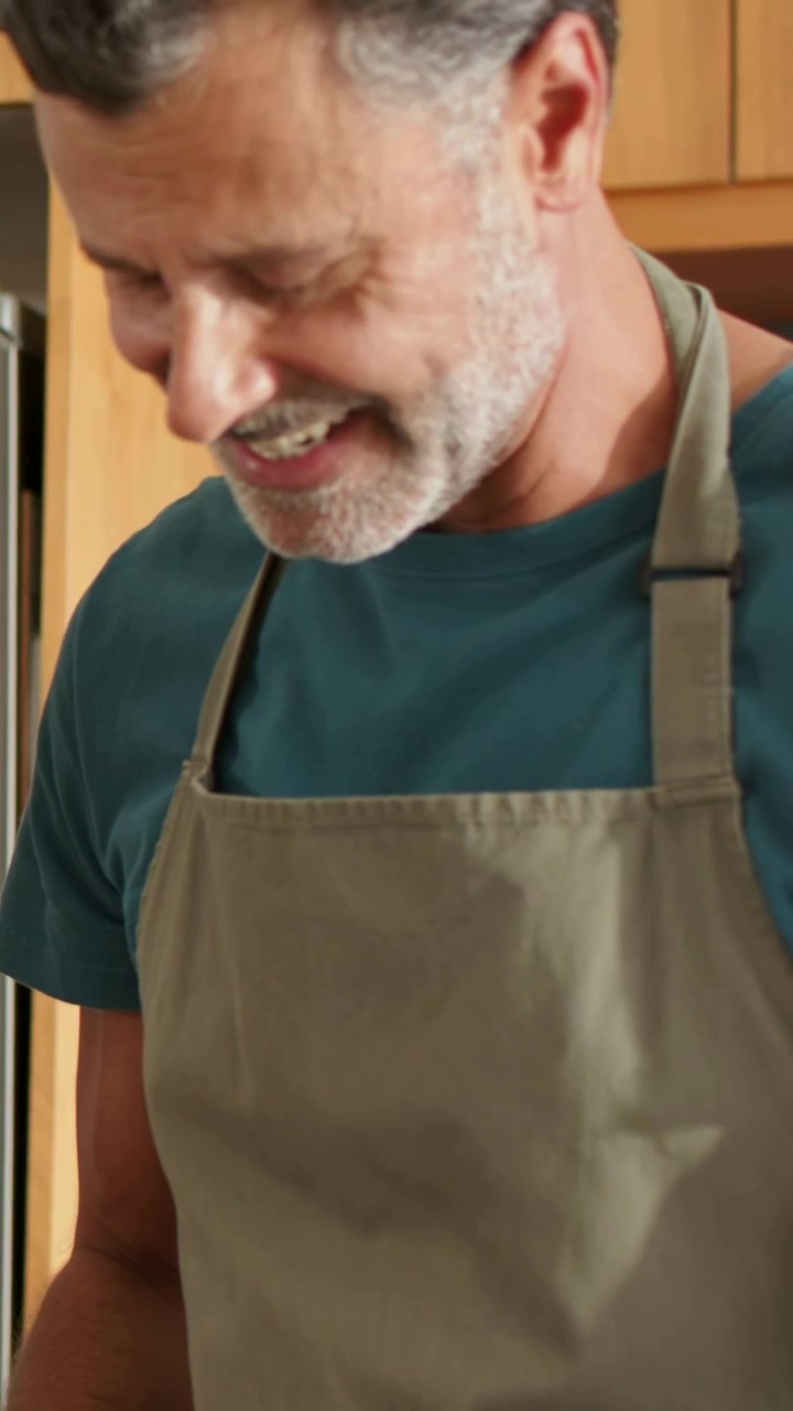 Video vertical: Hombre cocinando buscando algo en la cocina después de una llamada fuera de pantalla, con delantal verde oliva y reloj inteligente.