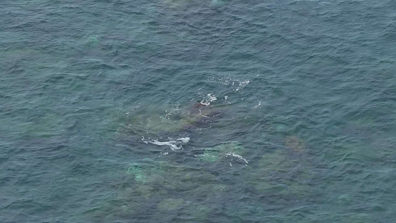Aerial view over small rock submerged reef with ocean waves washing over making white foam