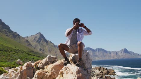 Afroamerikanischer-Mann-Beim-Wandern,-Sitzend-Auf-Einem-Felsen-Und-Fotografieren-An-Der-Küste