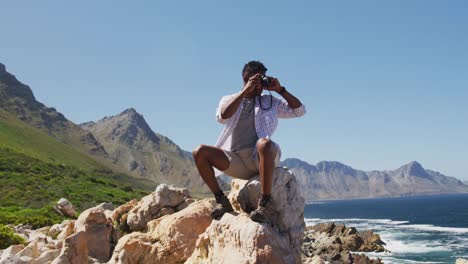 african american man hiking sitting on rock taking photos by the coast