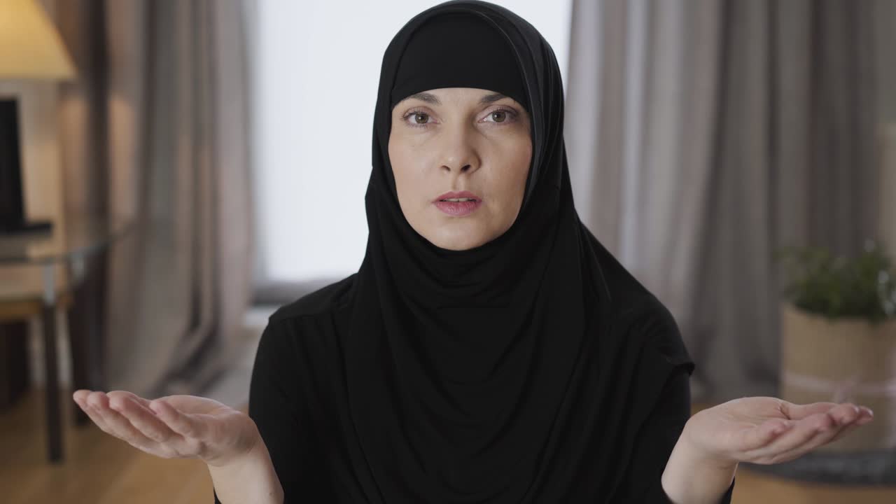 Close-up portrait of beautiful Muslim woman sitting with raised hands, looking up and talking. Young lady in traditional hijab praying at home. Religion, traditional culture.