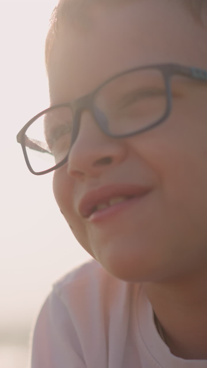 Close-up shot of a young boy wearing a white shirt and glasses, holding and happily eating a red apple in a sunlit field. The scene captures the joy of simple pleasures in nature