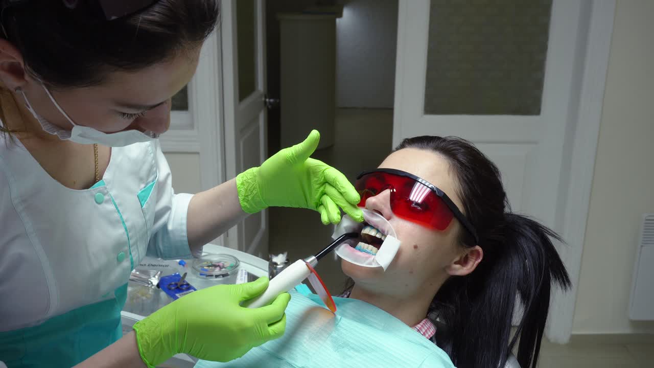Young woman with an expander in mouth and red protective glasses getting UV whitening at the dentist's office by an ultra violet machine. Shot in 4k