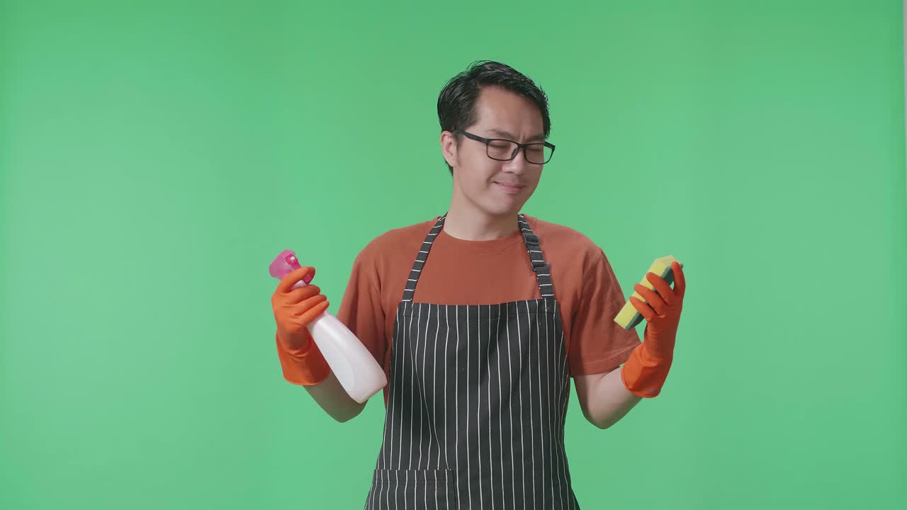 녹색 스크린 스튜디오 에 서 있는 동안 스프레이 와 주방 스폰지 를 보여 주는 에프론 을 입은 미소 짓는 아시아 남자 집주인