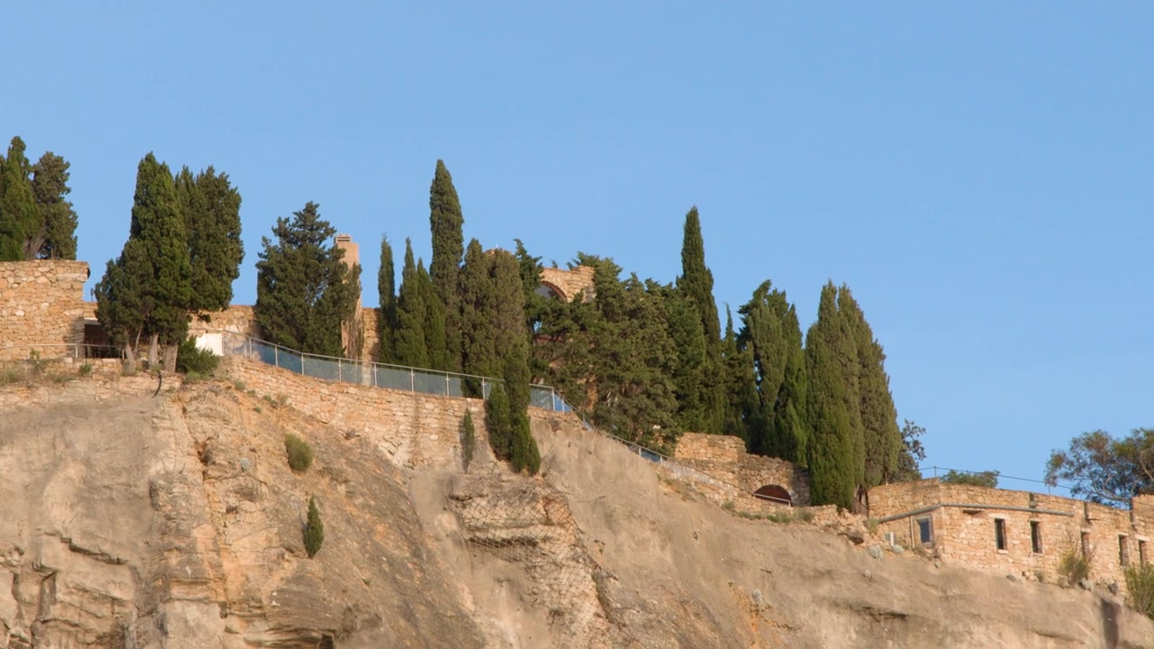 Inquadratura ampia di antiche mura di un castello in pietra in cima a scogliere rocciose sotto un cielo azzurro limpido.