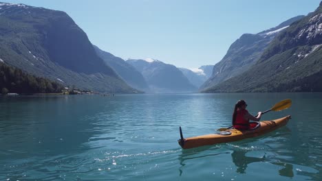 woman kayaking glacial green freshwater lake in loen - beautiful mountain landscape at sunny day - camera moving sideways close to water with kayak moving away into center of frame - norway