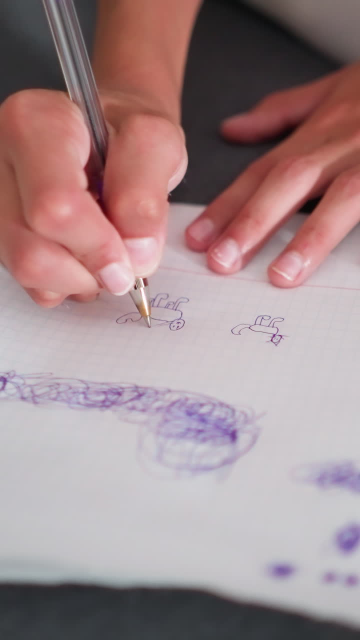 Primer plano de un niño sentado con las piernas cruzadas en un sofá dibujando garabatos creativos con un bolígrafo sobre un papel apoyado en una superficie suave, mostrando una concentración relajada y el movimiento de la mano en un ambiente hogareño informal.