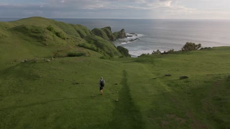 Sobrevuelo-Aéreo-Que-Muestra-A-Un-Hombre-Caminando-Sobre-Un-Paisaje-De-Colinas-Cubiertas-De-Maleza-Con-Un-Hermoso-Océano-De-Fondo