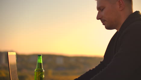 a male freelance programmer sits on a skyscraper roof with a laptop and beer typing code on a keyboard during sunset. remote work