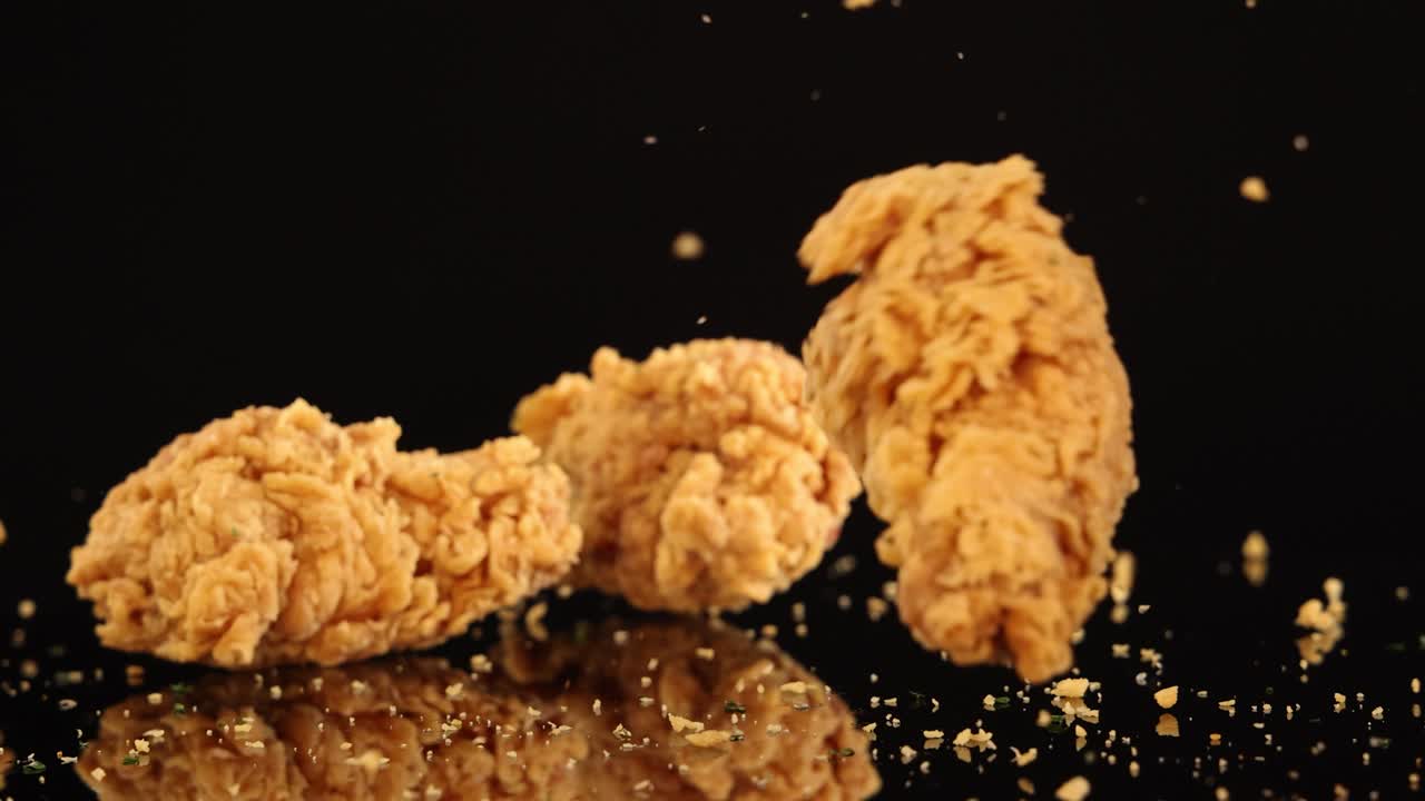 Golden fried chicken pieces fall and bounce on a glossy black surface, scattering crumbs. Studio lighting highlights texture, with slow-motion effect and dramatic mood