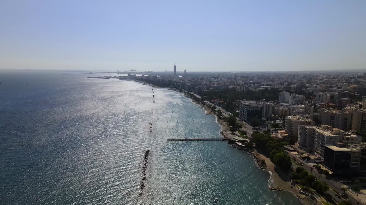 Drone video captures expansive shoreline of Durrës, Albania, with shimmering sea, breakwaters, towering skyscraper, and urban landscape under clear sky. Stunning aerial footage