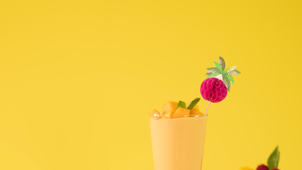 Una mano inserisce una cannuccia a righe in un frullato tropicale al mango tra frutta fresca su sfondo giallo