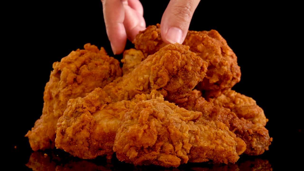 A hand reaches into a pile of golden, spicy fried chicken pieces, picking one up in slow motion. Dramatic lighting and black background emphasize texture