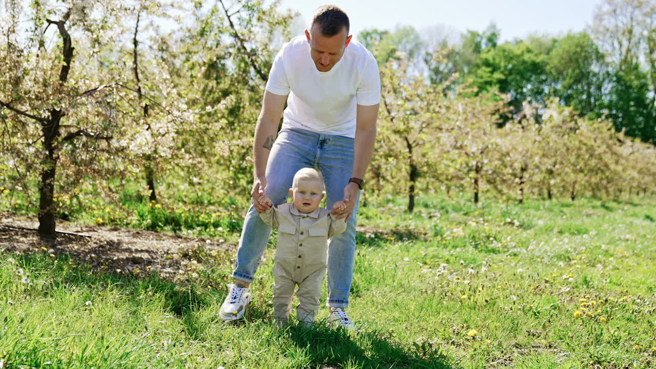 Caucasian man wearing a t-shirt and jeans stands leaned over his baby. Father is supporting his son learning to walk outdoors.