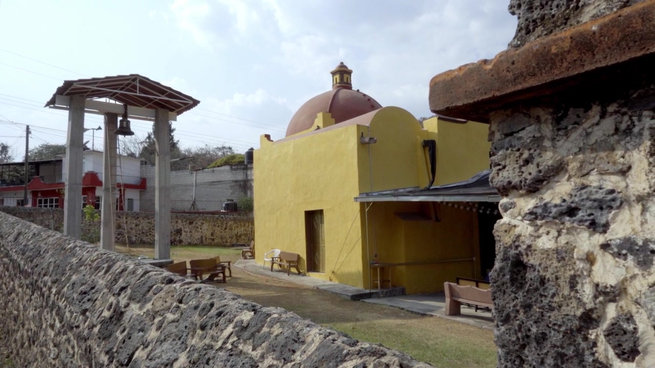 Piccola chiesa gialla con cupola e facciata semplice, struttura campanaria nelle vicinanze e muro in pietra in primo piano, ambientazione rurale con panchine e spazio aperto alla luce del giorno.