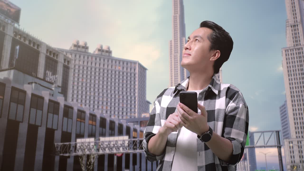 hombre asiático independiente hablando en teléfono inteligente y mirando a su alrededor mientras está de pie frente a los edificios trabajando en una ciudad