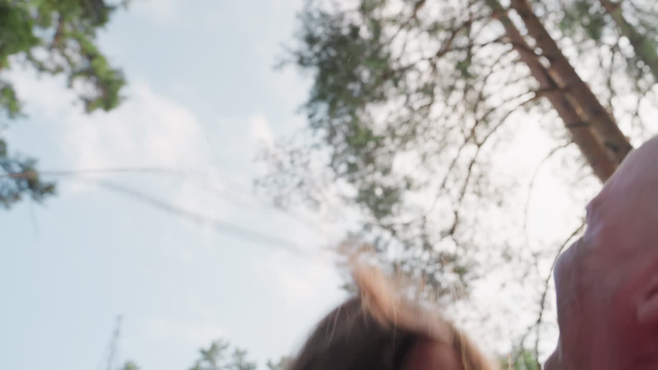 Sky view showing excited teenage girl laughing as dad throws her up and catches her during joyful family playtime in nature, surrounded by bright sunlight, green trees, and warm atmosphere of fun