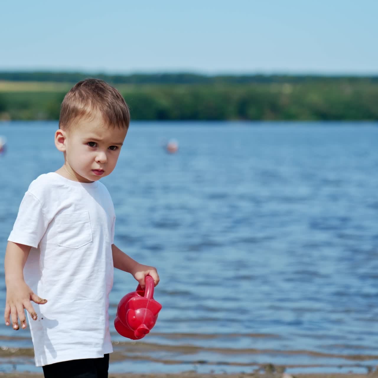 Caucasian toddler walks by the beach holding a watering can. Kid walks by the sand carefully