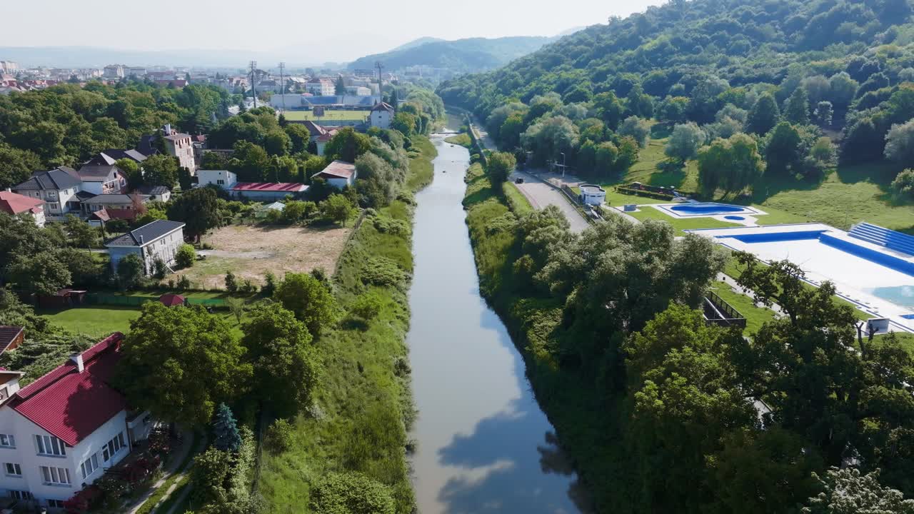 Bidikan drone udara lebar perlahan terbang maju di atas kanal sungai lurus yang membagi lingkungan perumahan dan area hutan hijau subur, dengan jembatan melengkung melintasi air