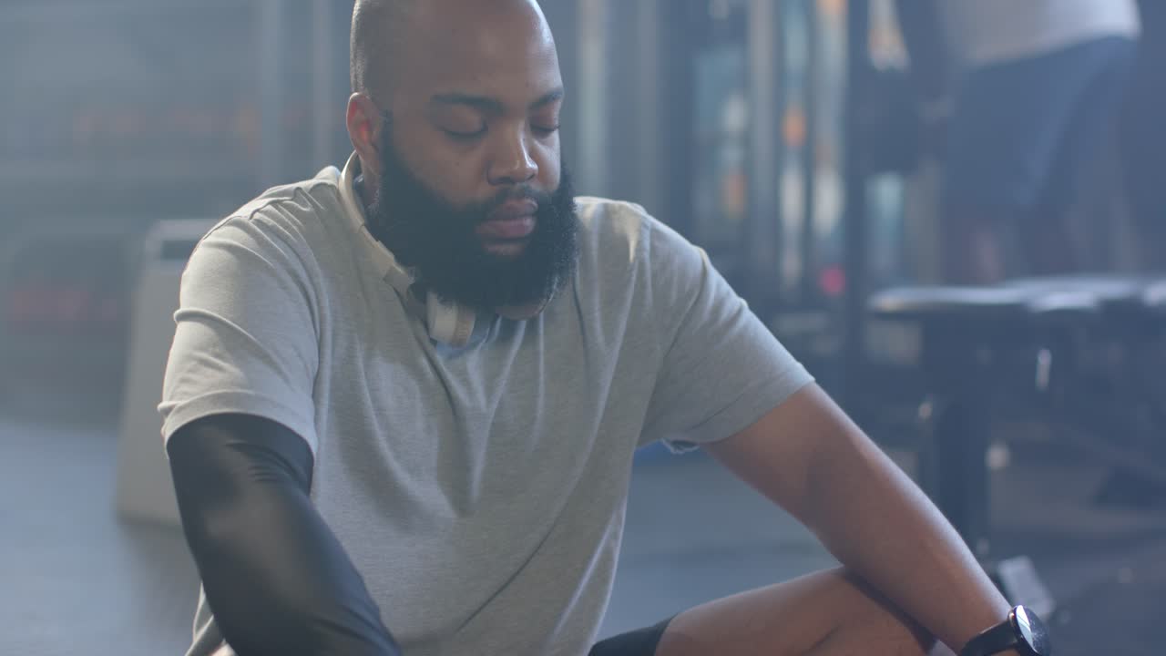 Pria Afrika berbusana olahraga duduk di gym menyesuaikan lengan, memasang headphone, bernapas untuk memulihkan diri
