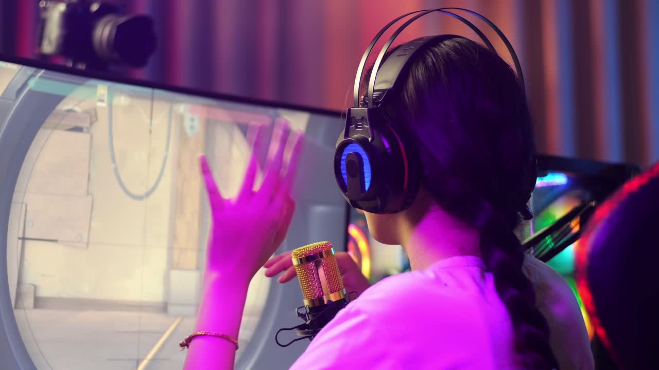 vista de atrás de una chica asiática jugando a un juego a través de la red en una computadora personal, hablando con los participantes en el micrófono. videojuego de transmisión en vivo, escritorio iluminado por luz de tira de led rgb