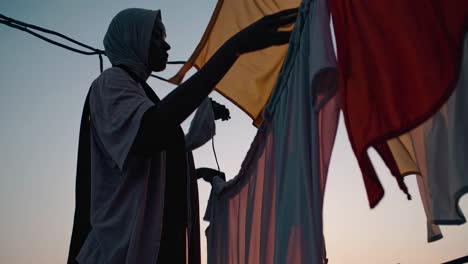 woman wearing a hijab hangs laundry on a clothesline on a rooftop at sunset, creating a serene and domestic scene against the colorful sky