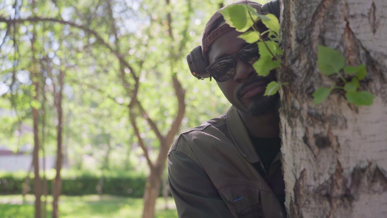 Close up of man wearing headphones standing in outdoor park setting with trees and greenery. subject remains mostly stationary framed against natural background. Soft daylight illuminates