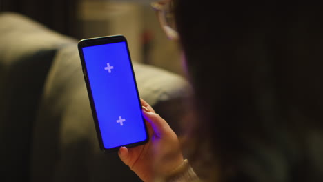 close up over the shoulder shot of woman spending evening at home sitting on sofa looking at blue screen mobile phone