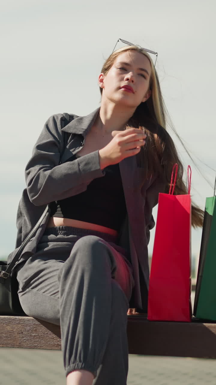 mujer sentada en un banco al aire libre con tres bolsas de compras de colores a su lado, cruzando las piernas y ajustando el cabello mientras posa con estilo en un día soleado, postes eléctricos visibles en el fondo