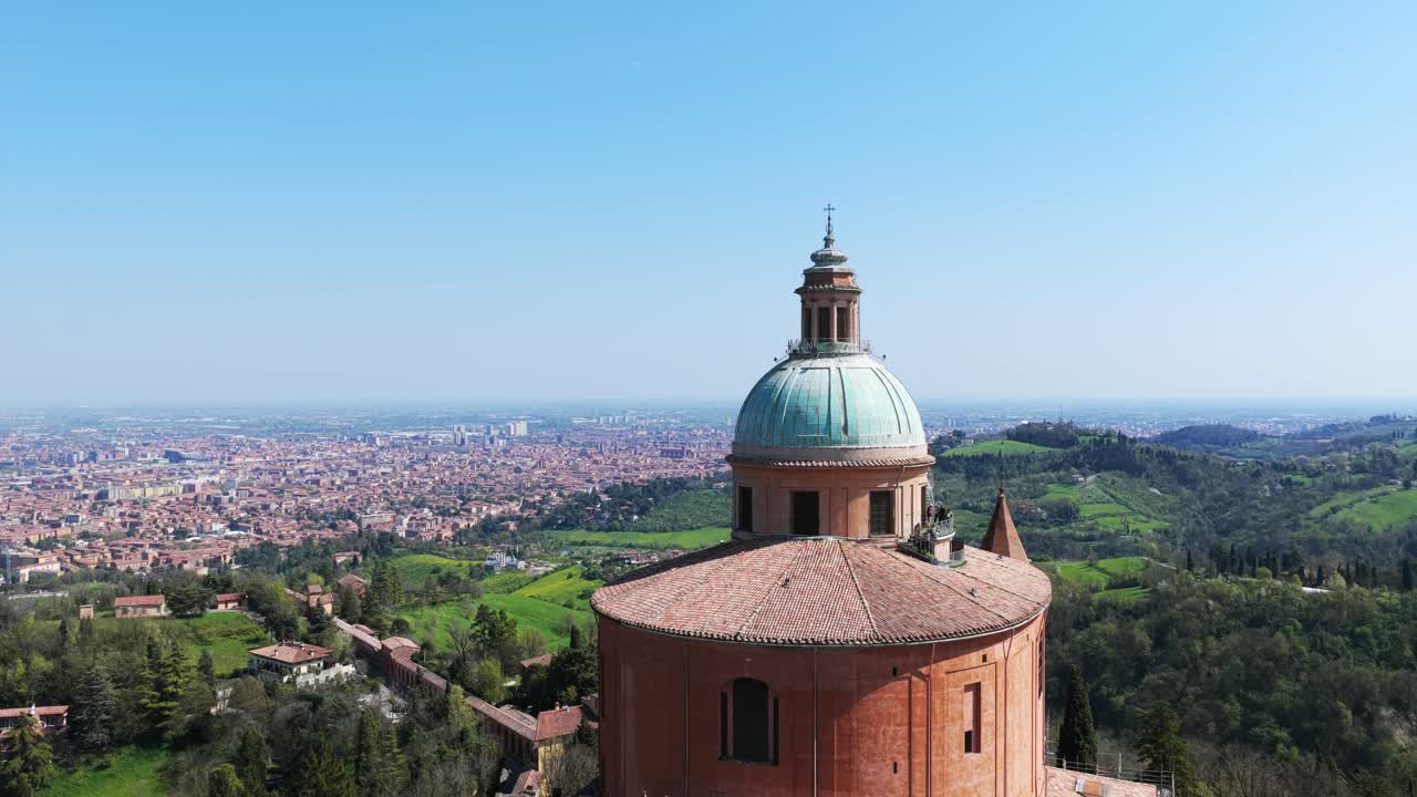 Dolly backward aerial shot of the Sanctuary of San Luca in Bologna, revealing the iconic dome and Baroque architecture with the sprawling cityscape and green hills in the background under a clear sky