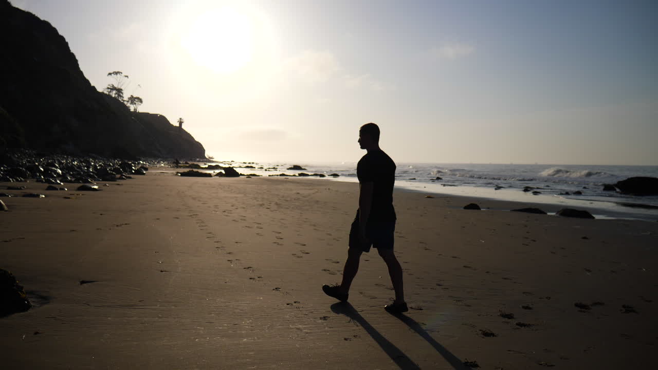 un hombre en silueta caminando por la playa al amanecer con olas rompiendo en la costa en santa barbara, california a cámara lenta