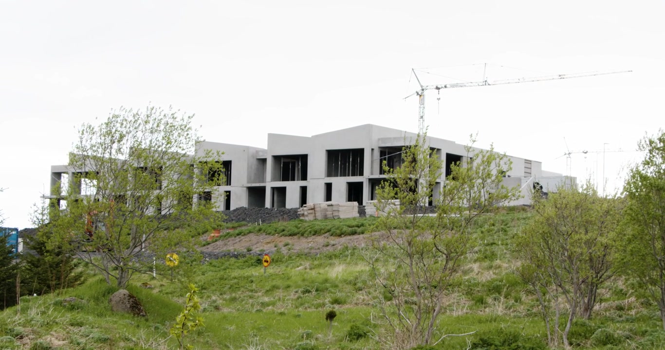 En stor, moderne betongbygning under oppføring ligger på en grønn åsside med en høy kran i bakgrunnen. Forgrunnen er fylt med trær og frodig gress