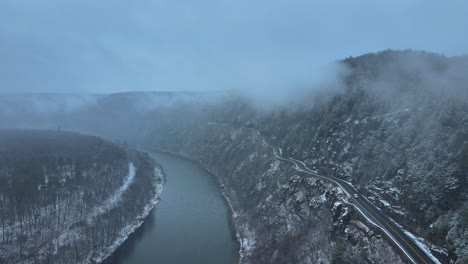 aerial footage of a snowy, scenic byway, winding mountain valley road during a snowstorm with pine trees, a river, mountain highway, rocky cliffs, and forests during winter on a cold, blue day