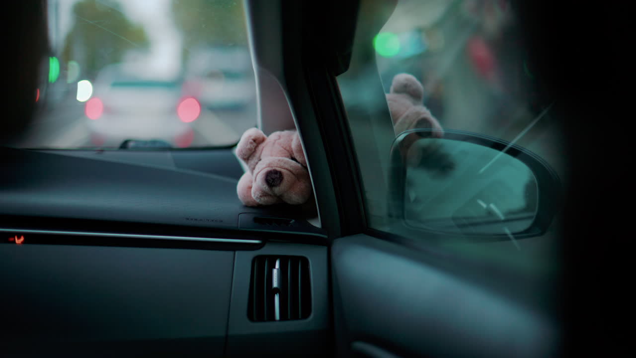 A small stuffed toy rests on a car dashboard as blurred city lights pass by through the windshield