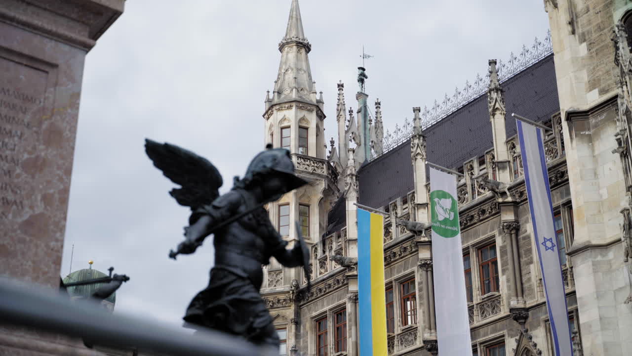 A silhouette of an angel statue in front of the New Town Hall in Munich, Germany. Gothic architecture with Ukrainian, Israeli, and environmental flags. Captured on a cloudy day.