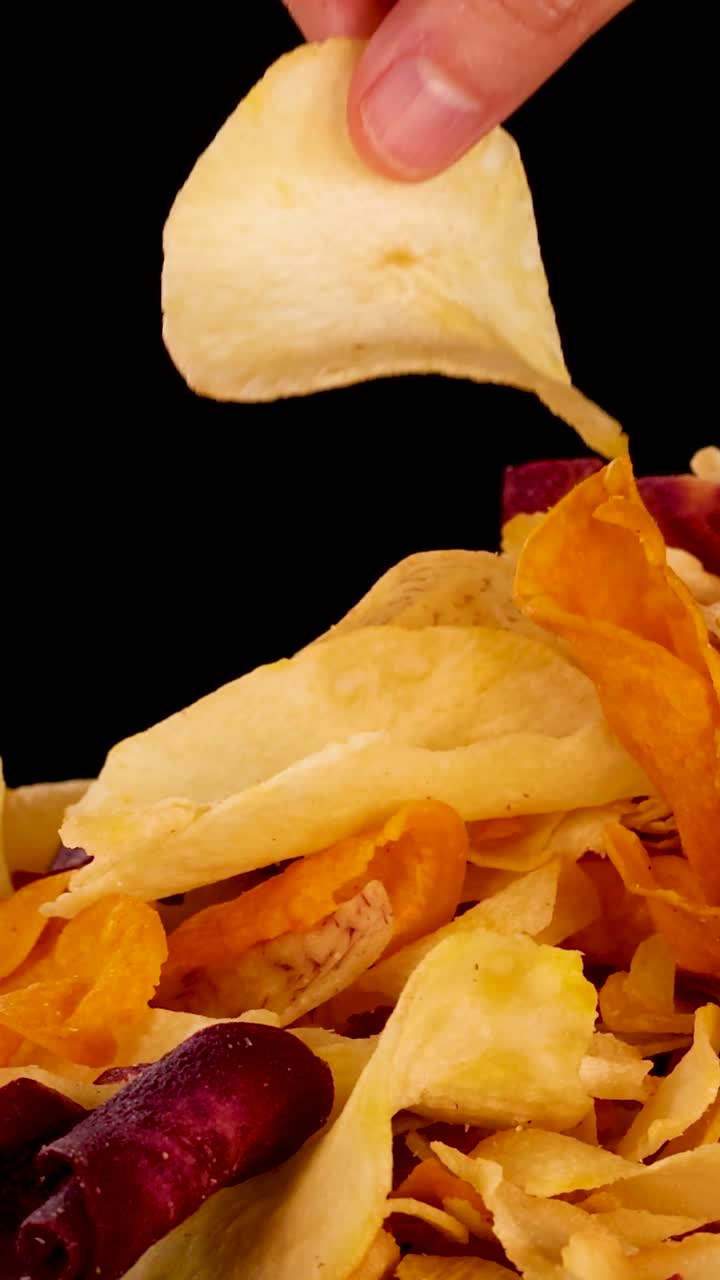 Close-up of a hand selecting colorful vegetable chips from a pile against a black background