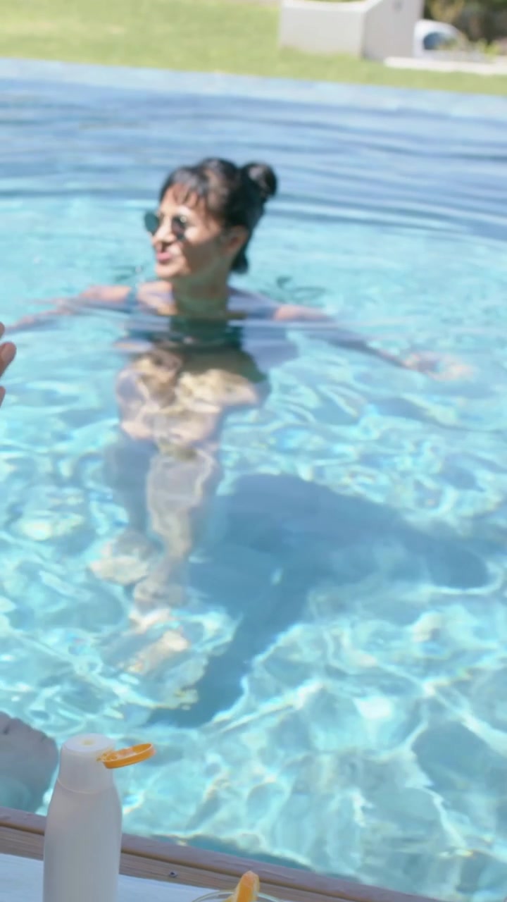 Video verticale: Donna sorridente in bikini che vede una mano offrire una bottiglia e si protende per prenderla a bordo piscina.