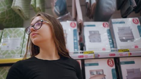 close-up of female shopper walking past store shelf filled with bedding products, she looks around, observing fabric designs and textile options. home decor, linens, and household essentials