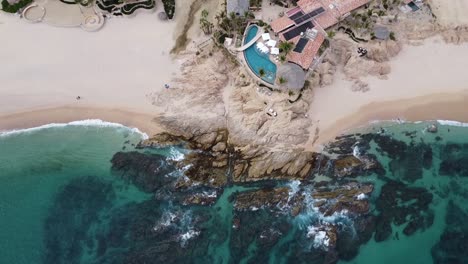 aerial birdeye view of a gorgeous coastline with turquoise sea while the waves break on the rocks right on the beach with a beautiful villa with swimming pool on a trip through mexico at chileno beach
