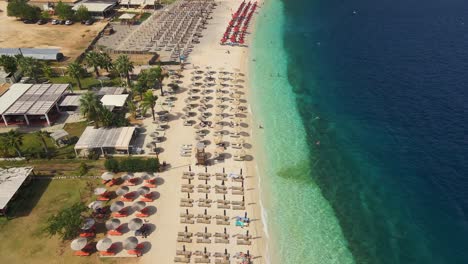 Parallax-drone-shot-moving-along-Myrtos-beach,-full-of-beach-chairs-in-Greece-during-summer